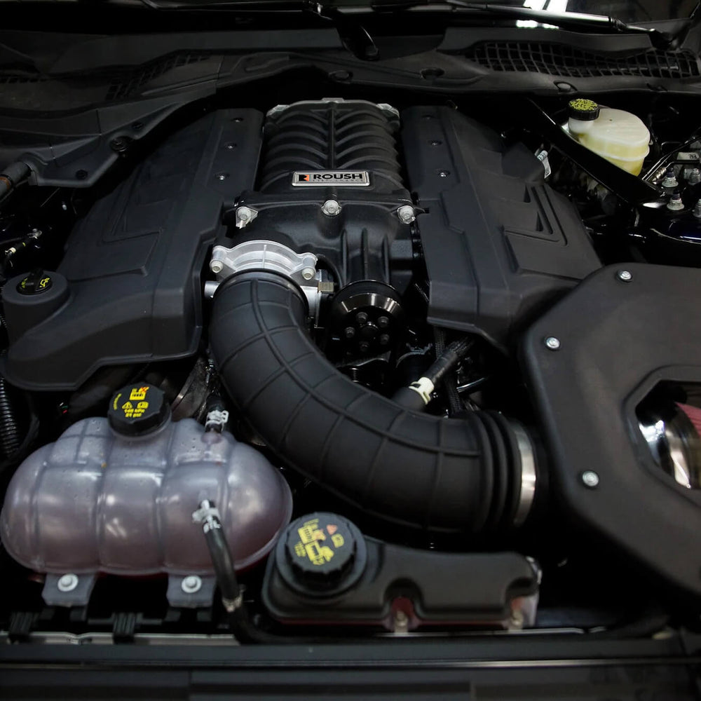 An engine compartment of a Mustang GT featuring a prominent Roush supercharger, various hoses, and interconnected components visible, delivering an impressive 750 horsepower.