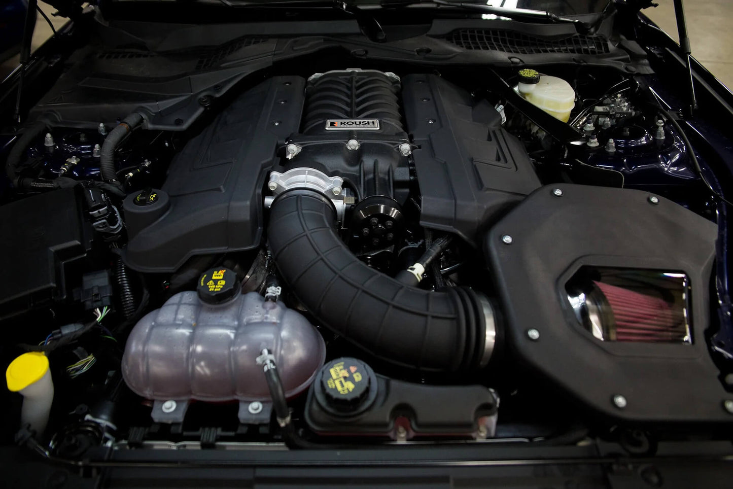An engine compartment of a Mustang GT featuring a prominent Roush supercharger, various hoses, and interconnected components visible, delivering an impressive 750 horsepower.