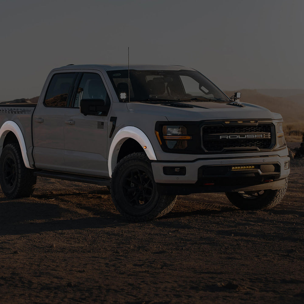 A white pickup truck, equipped with an F-150 fender flare kit, sits proudly on a dirt road in the desert landscape, under a clear sky with majestic mountains in the background.