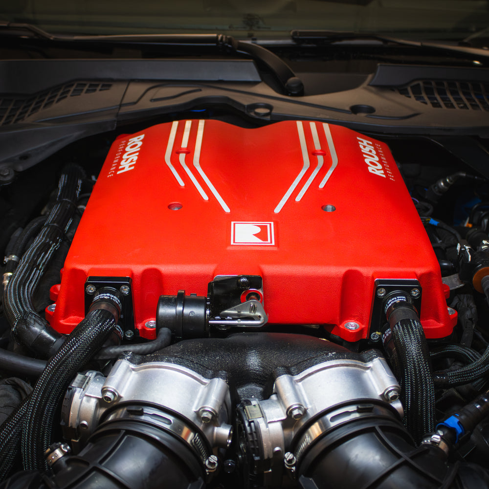 Close-up of a car engine with a striking red cover featuring "Roush" branding and white stripes. The TVS R2650 Supercharger is prominently displayed, boosting this Roush Mustang Supercharger to deliver an impressive 810 horsepower. Various components and wires add to its robust appearance.